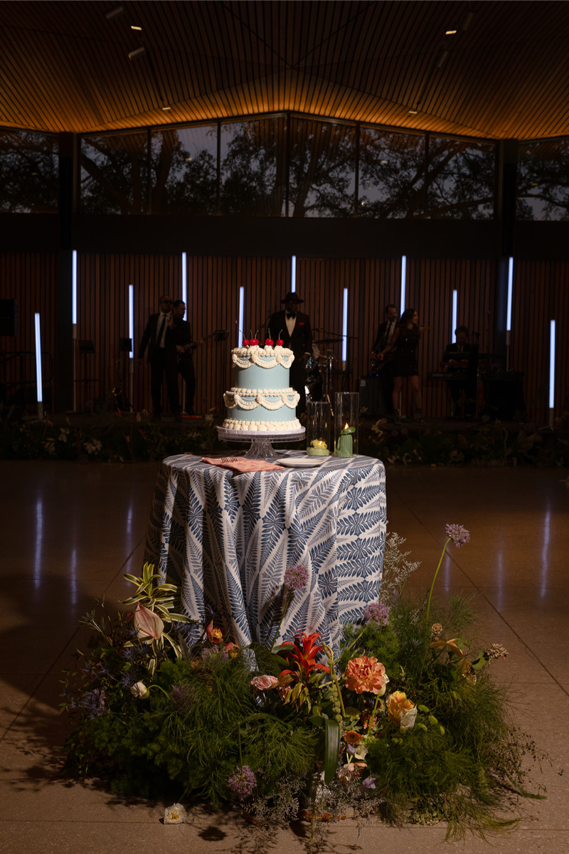 A tiered cake with blue and white icing sits on a patterned tablecloth, surrounded by flowers, with a band performing in the dimly lit background.