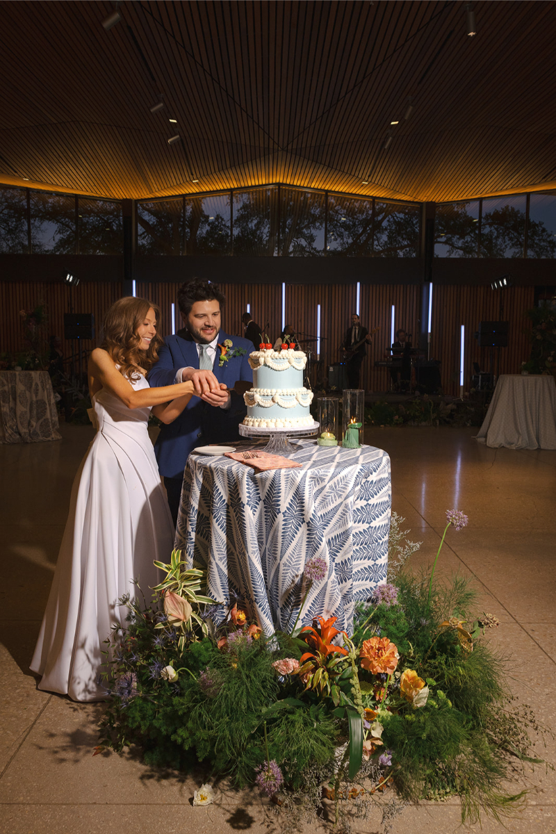 A bride and groom cut a tiered wedding cake together at a decorated table, surrounded by flowers, in a modern, warmly lit venue, representing cool wedding ideas the couple included.