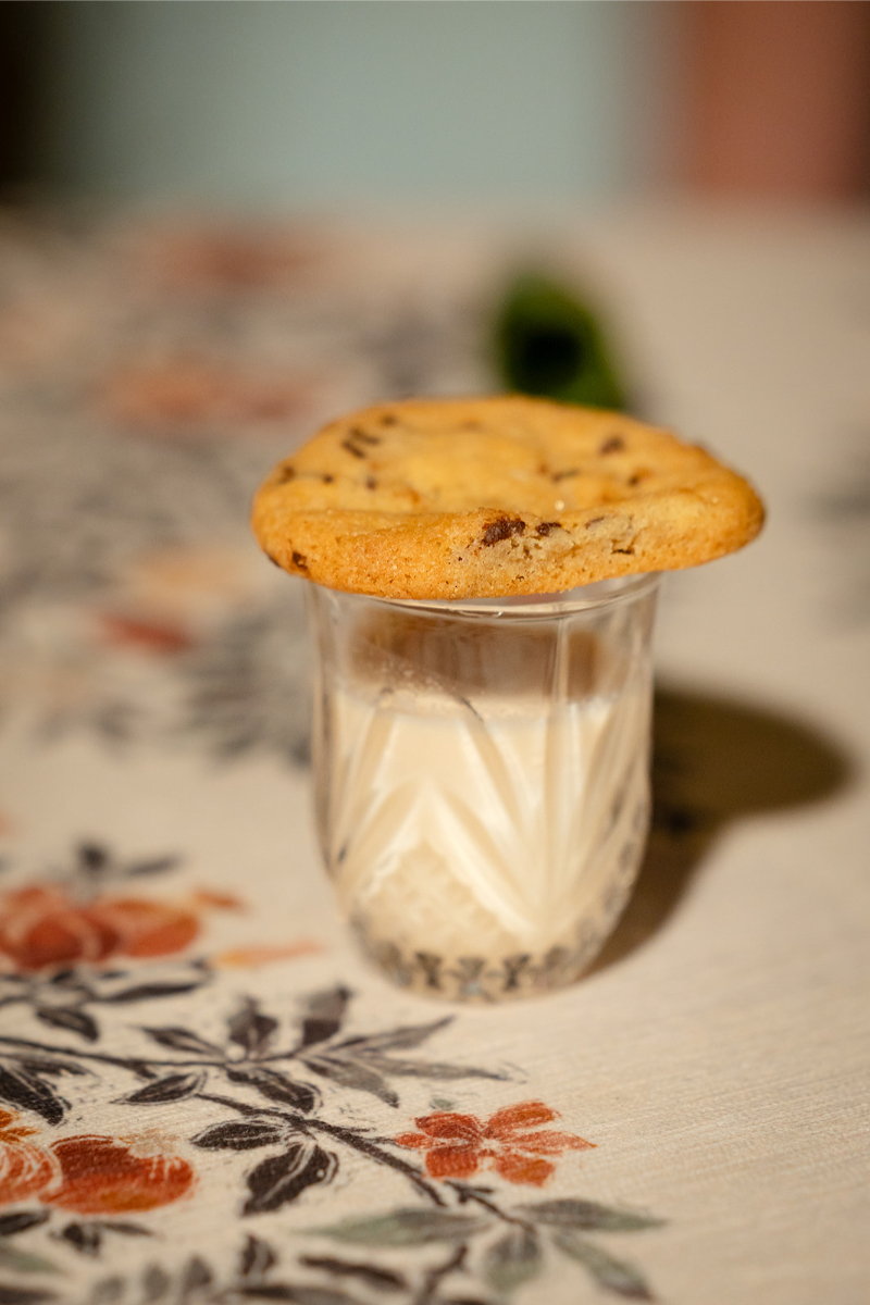 A chocolate chip cookie rests on top of a glass of milk, placed on a patterned tablecloth.