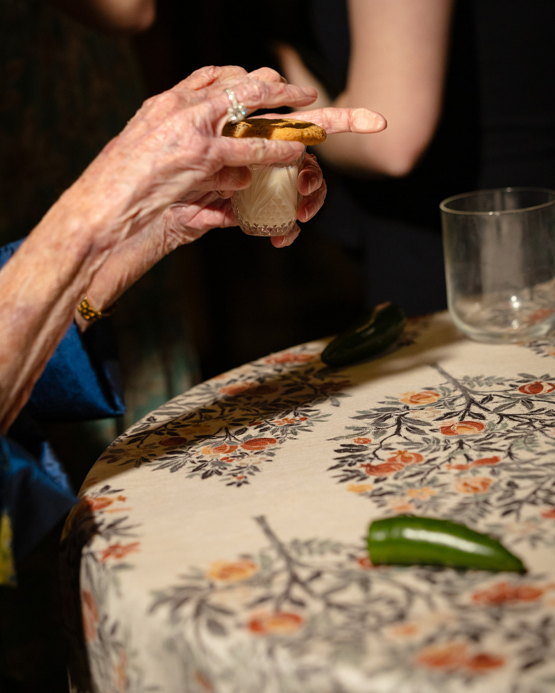 An elderly hand holds a shot glass with a cookie on top over a floral tablecloth, with a jalapeño and empty glass on the table.