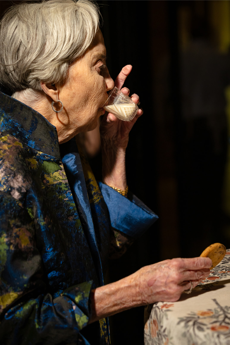 An elderly woman drinks from a small glass while holding a cookie, standing beside a table with a floral tablecloth.
