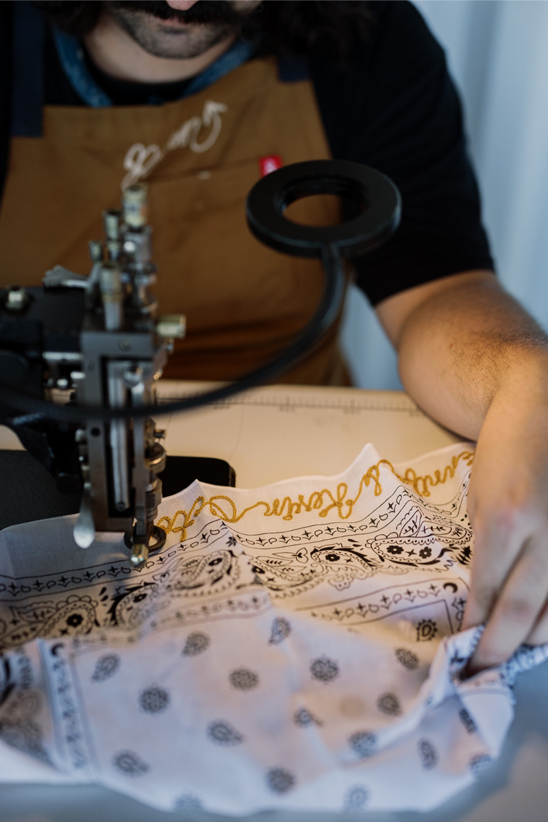 A person uses a sewing machine to embroider gold text onto a white patterned fabric while wearing a brown apron.
