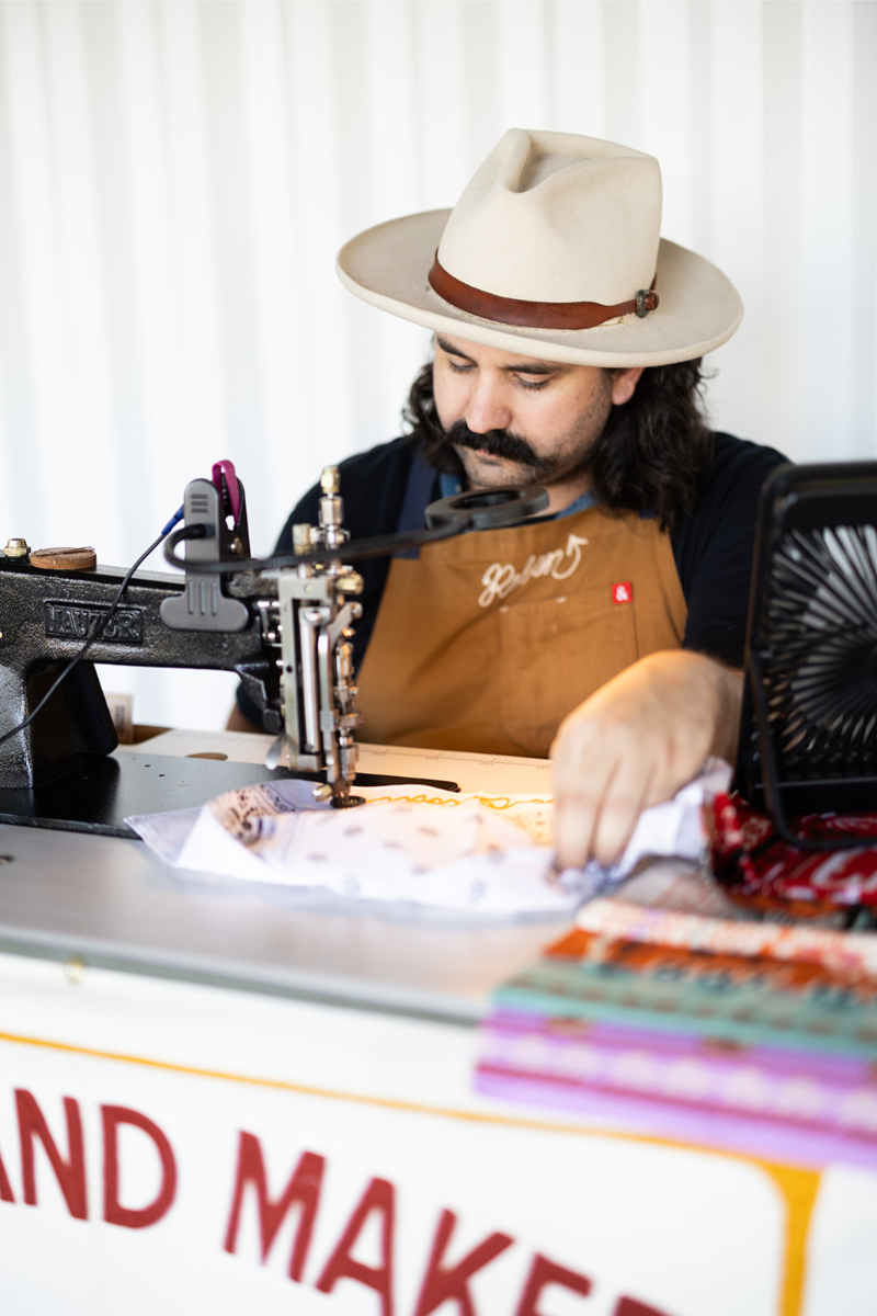 A person wearing a hat and apron operates an industrial sewing machine at a workbench with fabric and colorful cloths.