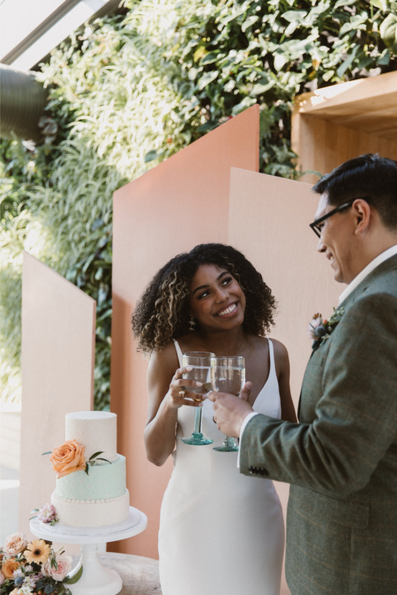 A bride and groom stand together smiling and holding glasses, with a two-tiered wedding cake on a stand beside them in a light-filled room with greenery, one of the top wedding weekend getaways in the US.