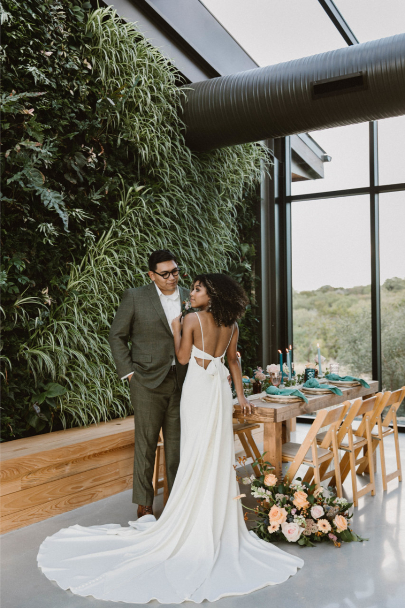 A bride in a white gown and a groom in a green suit stand together indoors next to a wooden table set for a meal, with a green plant wall and large windows in the background.