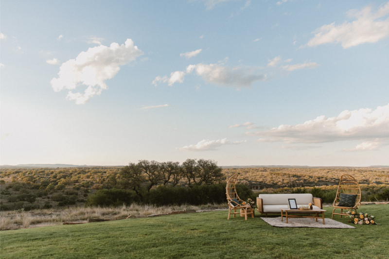 Outdoor seating area with a sofa, two chairs, a coffee table, and a rug on grass, set against a wide landscape under a partly cloudy sky.