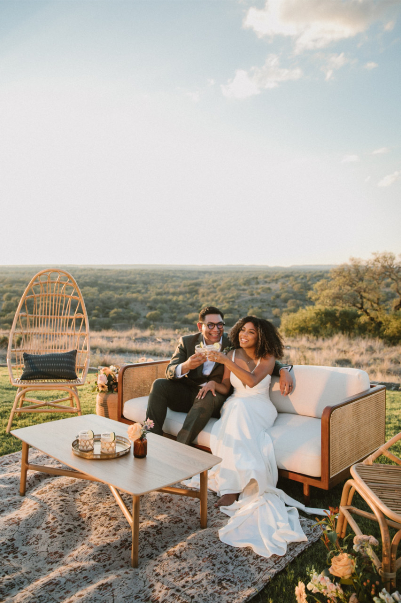 A bride and groom sit together on an outdoor sofa, smiling and holding drinks, with scenic hills and trees in the background.
