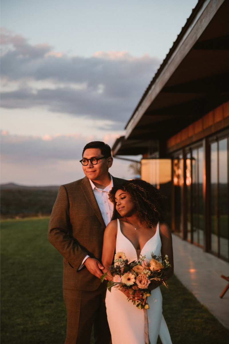 A couple in wedding attire stands close together outdoors at sunset; the groom wears a suit and glasses, and the bride holds a bouquet, with a modern building in the background, one of the top wedding weekend getaways in the US.