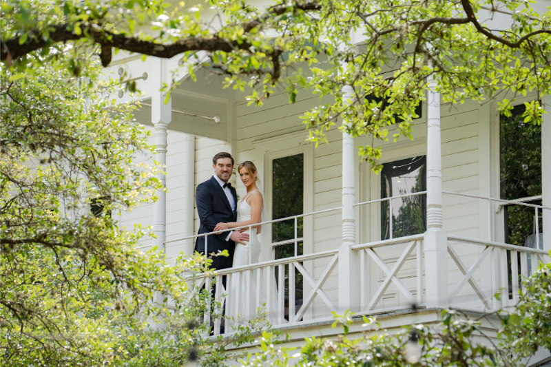 Wedding couple stand together on the balcony of a white wooden house surrounded by green trees