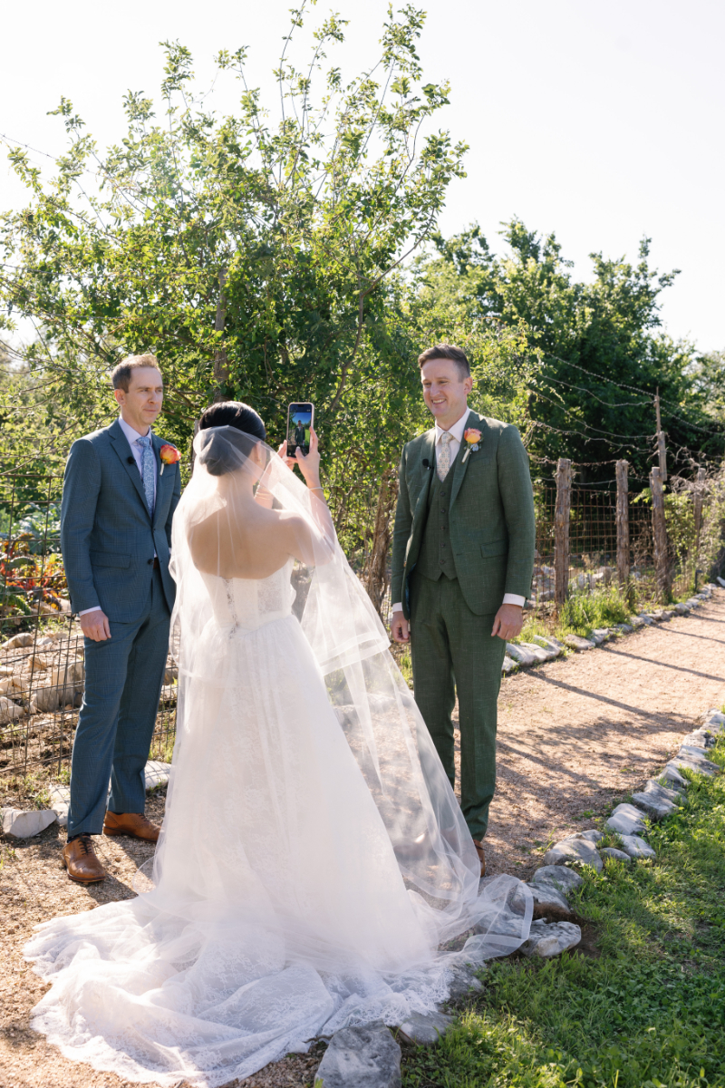 A bride in a white gown takes a photo of two men in suits standing outdoors on a garden path, with greenery and sunlight in the background.