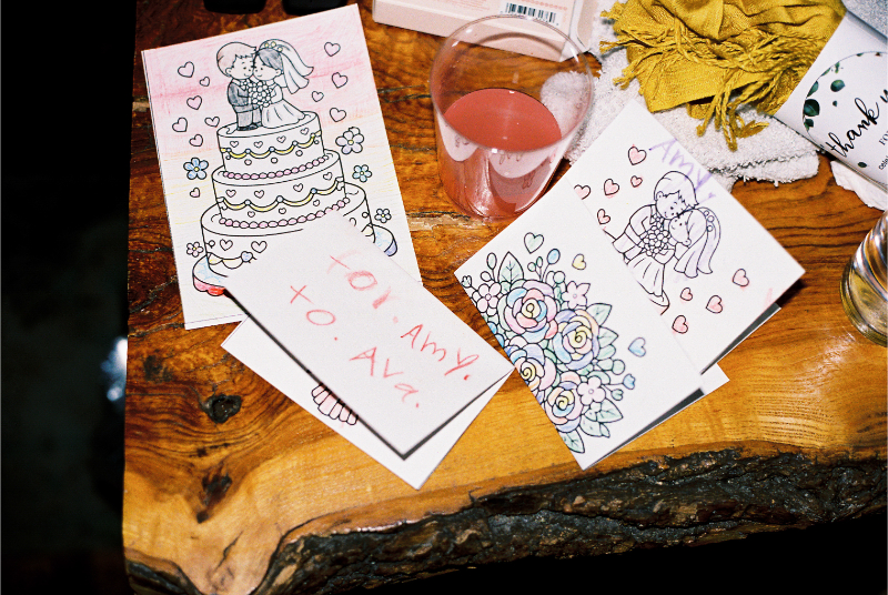 Coloring pages with wedding cake and angel designs, a pink drink, a handwritten card that says "To: Ava," and a yellow cloth rest on a wooden surface.