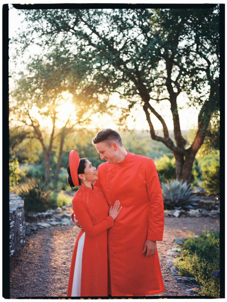 A couple in matching red traditional attire stands on a path outdoors at sunset, looking at each other and smiling, with trees in the background.