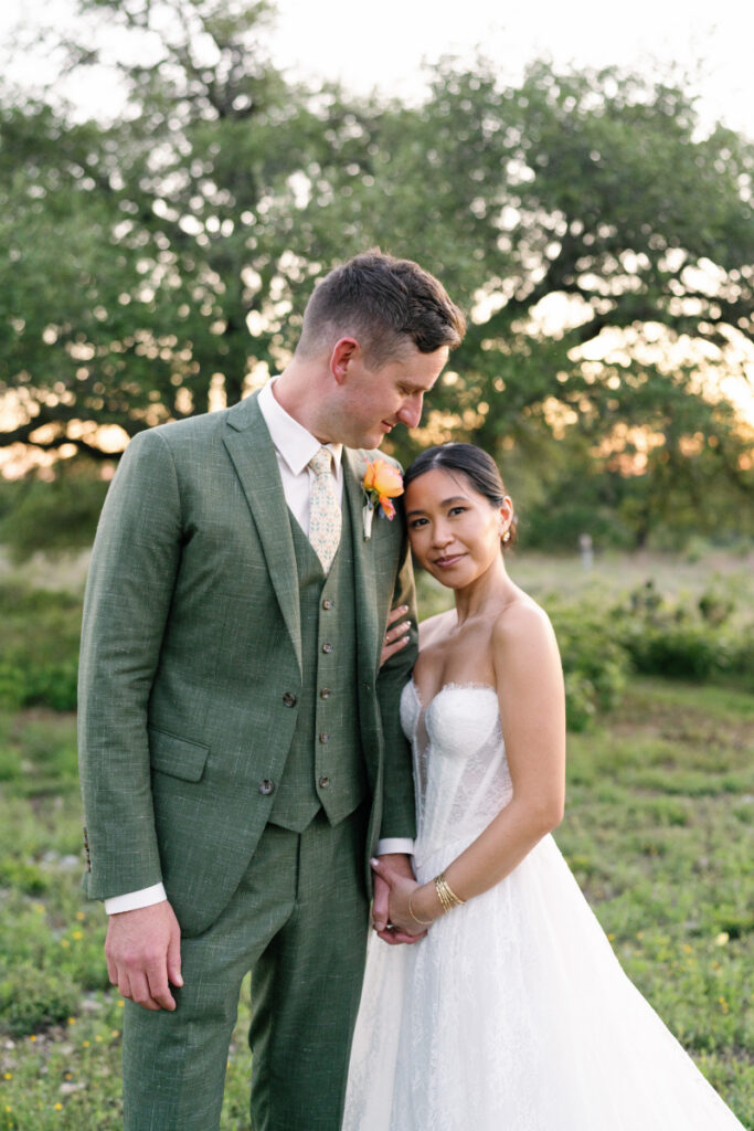 A bride in a white dress and groom in a green suit stand close together outdoors, holding hands, with trees and greenery in the background.