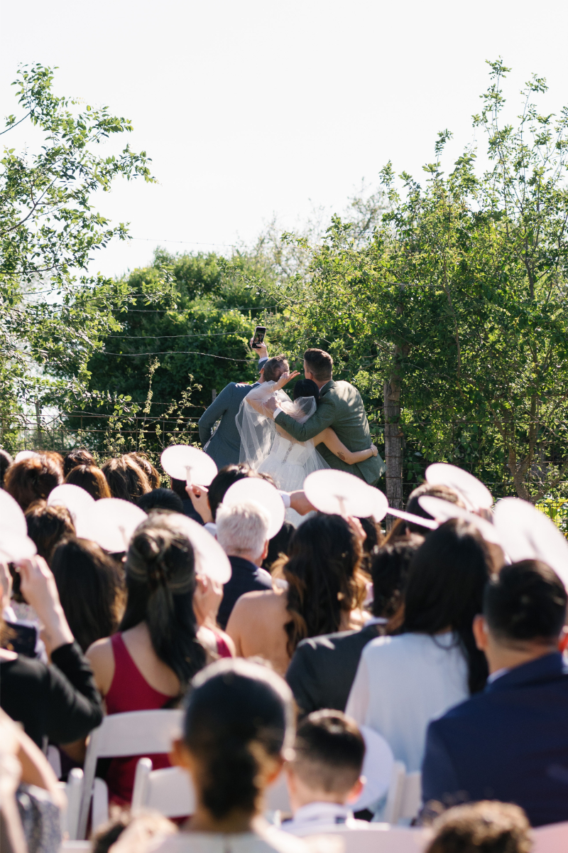 Outdoor wedding ceremony with a crowd seated facing a couple at the altar, surrounded by greenery, with two people standing beside the couple.