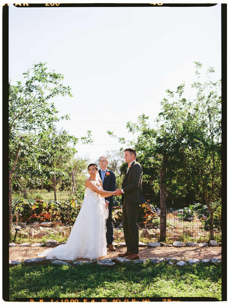 A bride and groom stand facing each other holding hands outdoors, with an officiant behind them, surrounded by greenery and a garden.