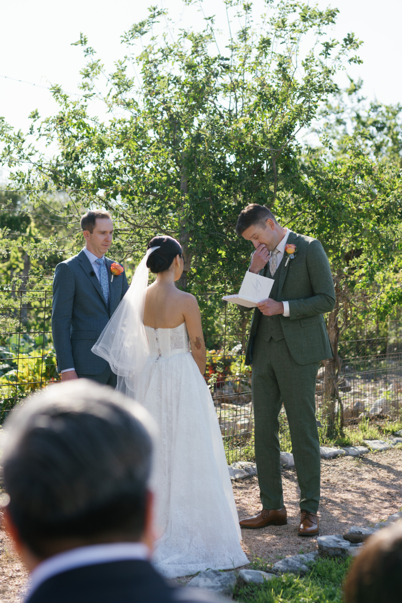 A bride and groom stand outdoors during their wedding ceremony. The groom reads from a paper, while an officiant stands nearby. Guests are partially visible in the foreground.
