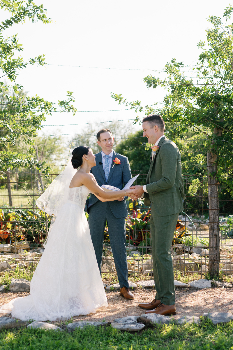 A bride and groom hold hands and smile at each other during an outdoor wedding ceremony with an officiant standing between them.