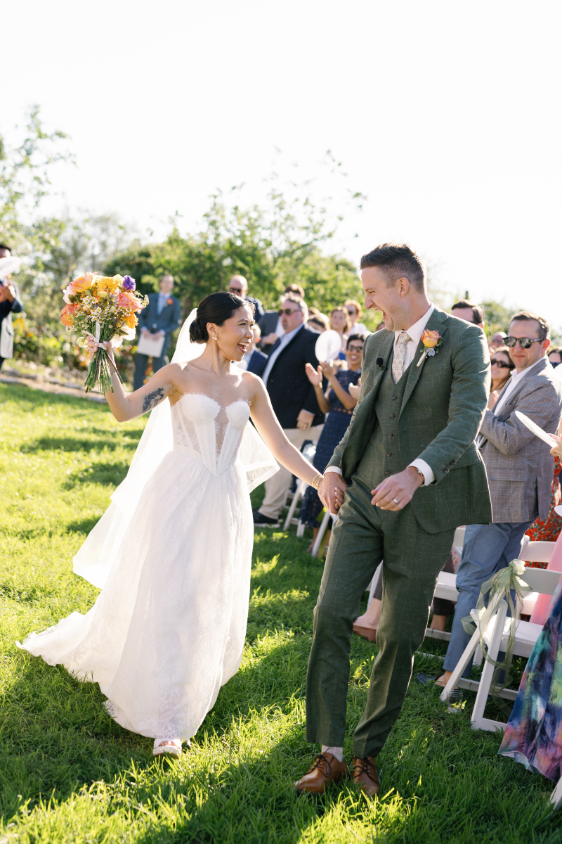 A bride and groom walk hand in hand outdoors after their wedding ceremony, smiling at each other as guests watch and applaud.