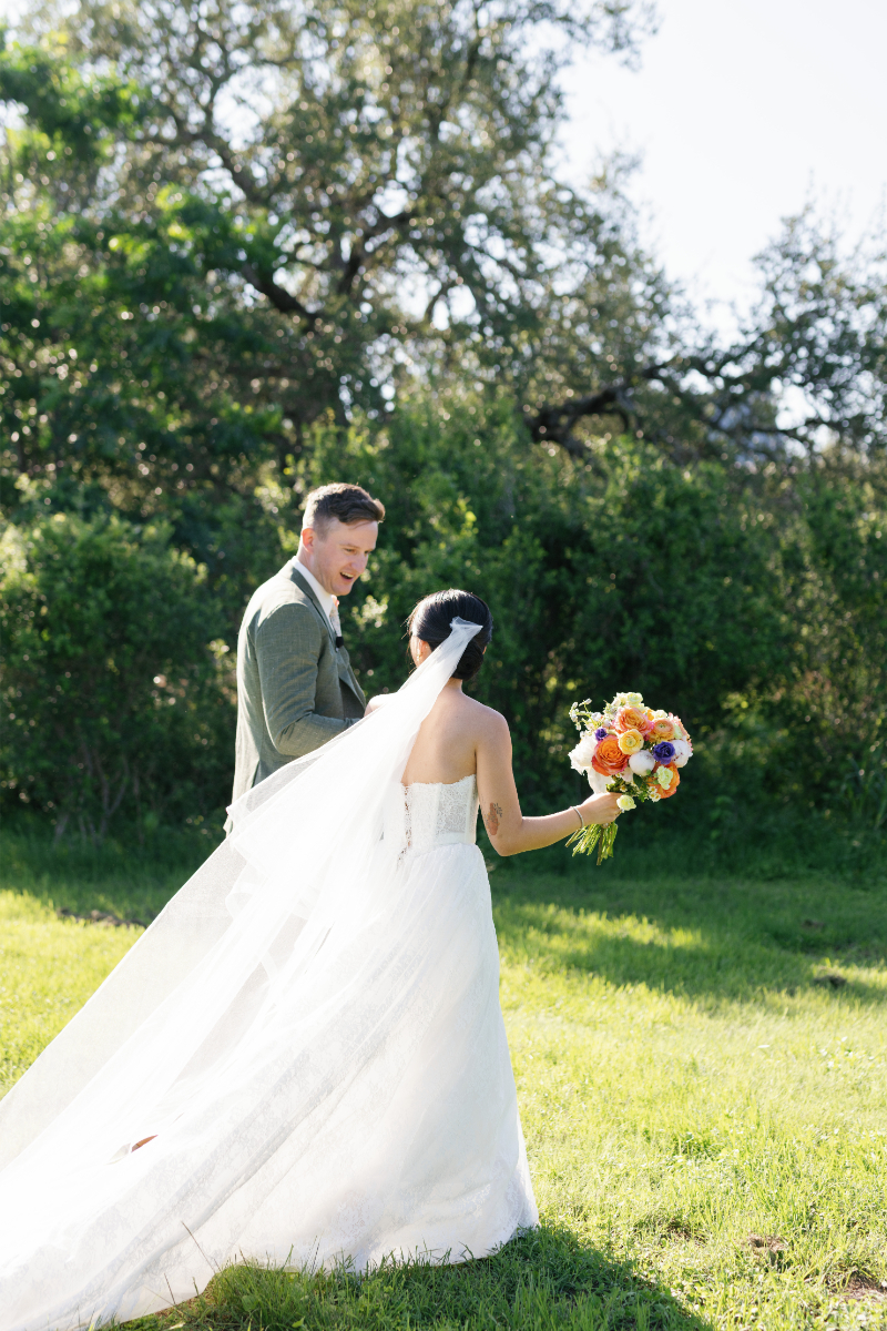 A bride in a white dress holding flowers stands beside a groom in a green suit outside on a sunny day, with trees and grass in the background.