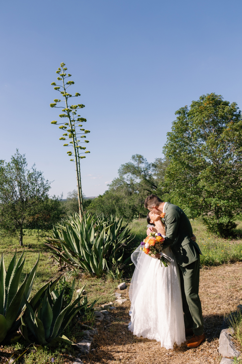 A bride and groom embrace outdoors on a garden path, surrounded by green plants and trees under a clear blue sky.