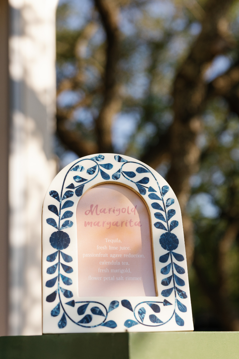 A decorative frame with blue leaf patterns displays a Marigold Margarita cocktail menu outdoors, with trees blurred in the background.