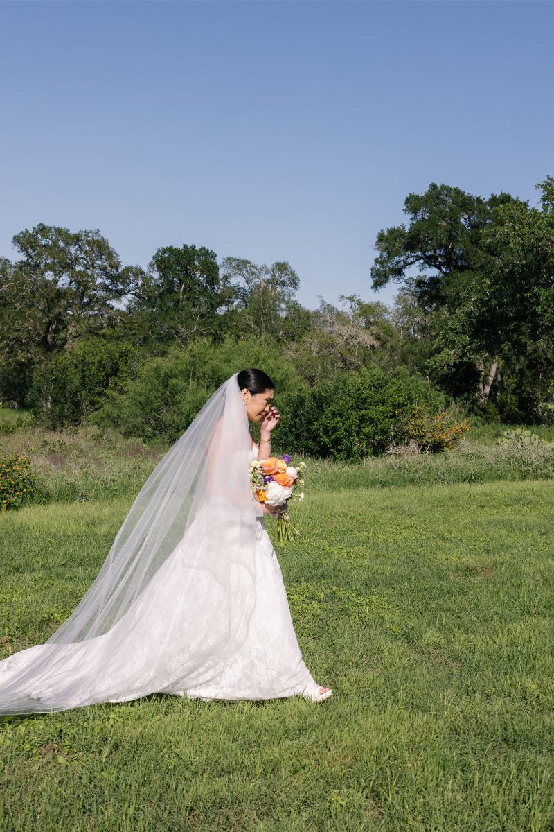 Bride in a white dress and veil holds a bouquet of flowers while walking on green grass with trees and a clear blue sky in the background.