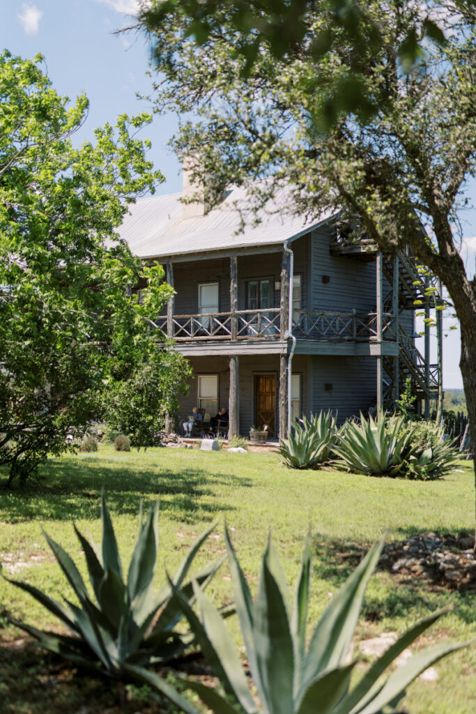 A two-story wooden house with a wraparound porch is surrounded by greenery, including agave plants and trees, under a partly cloudy sky.