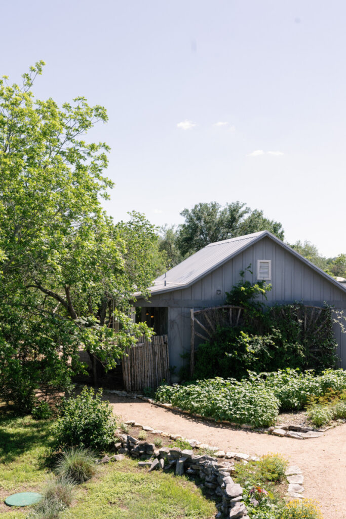A gray wooden house surrounded by lush greenery, a dirt path, and a stone border under a clear sky.
