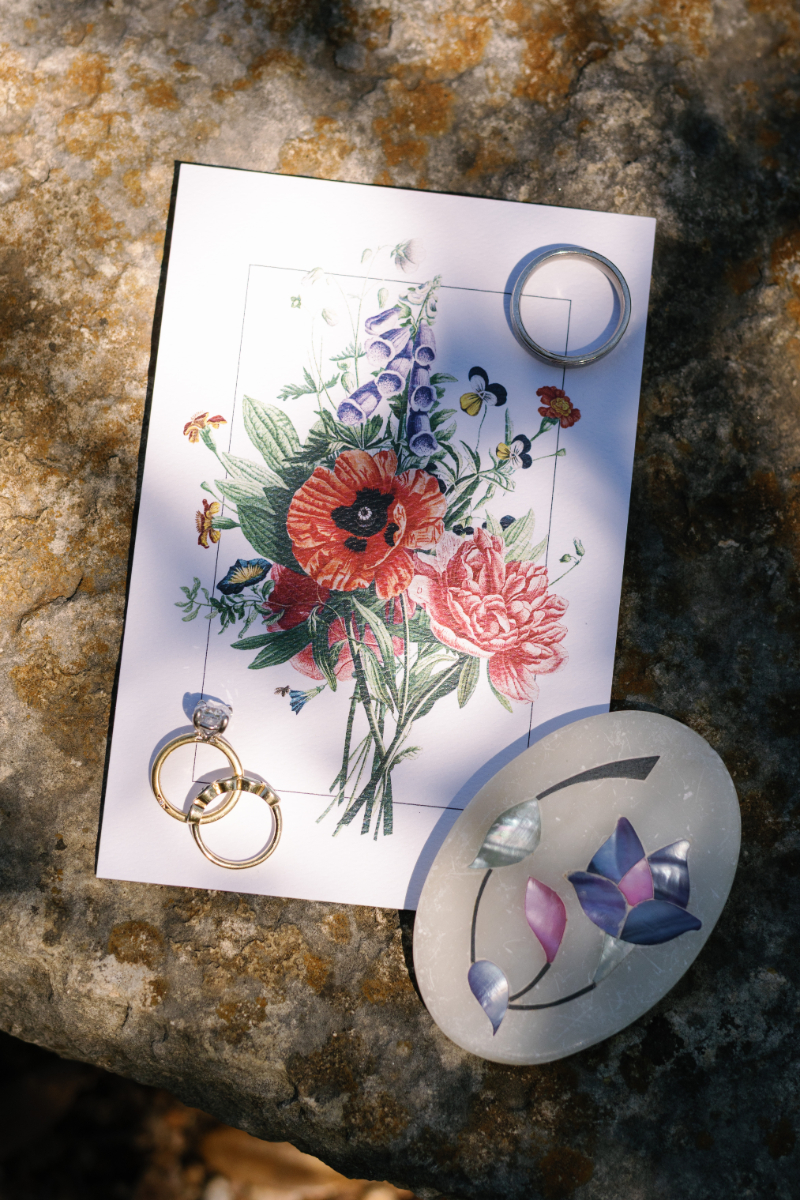 A floral illustration on paper is placed on a stone surface, surrounded by rings and oval stones in shades of purple, blue, and clear.