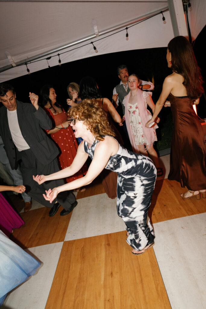 A group of people dance energetically under a tent at night; a woman in a black-and-white dress is in the foreground, bending her knees and smiling.