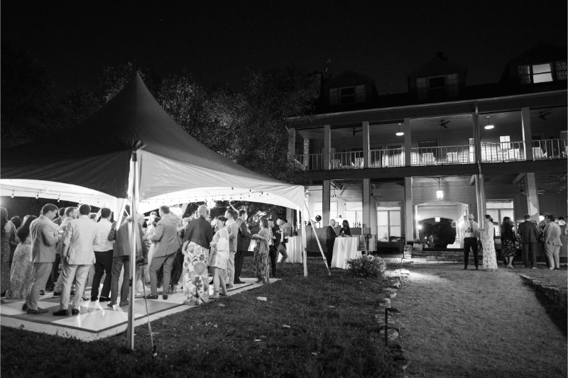 A group of people gather and dance under a tent outside a large, two-story house at night. The scene is lit with string lights and captured in black and white.