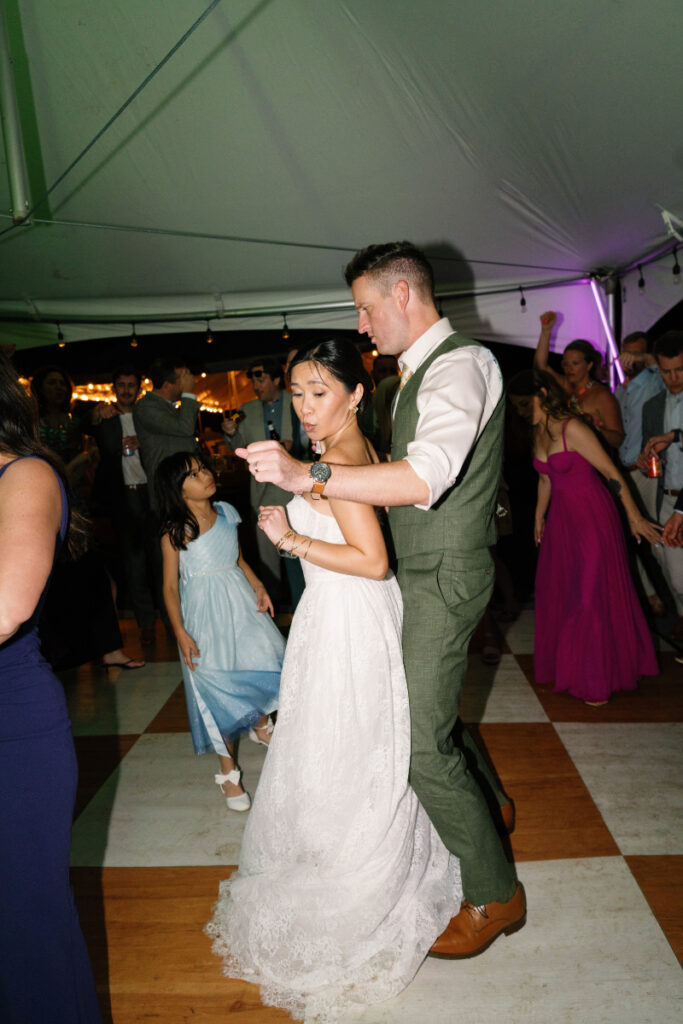 A bride and groom dance together at a wedding reception under a tent, with guests in colorful dresses dancing in the background.