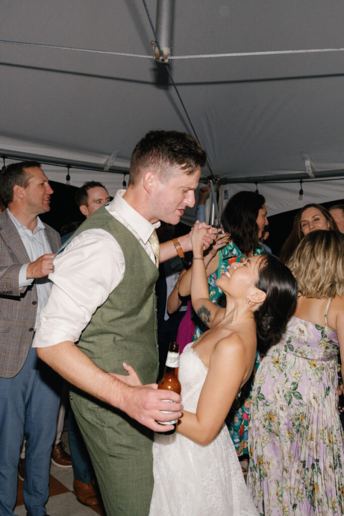 A man in a vest and a woman in a white dress dance together at a lively party, surrounded by other guests under a tent.