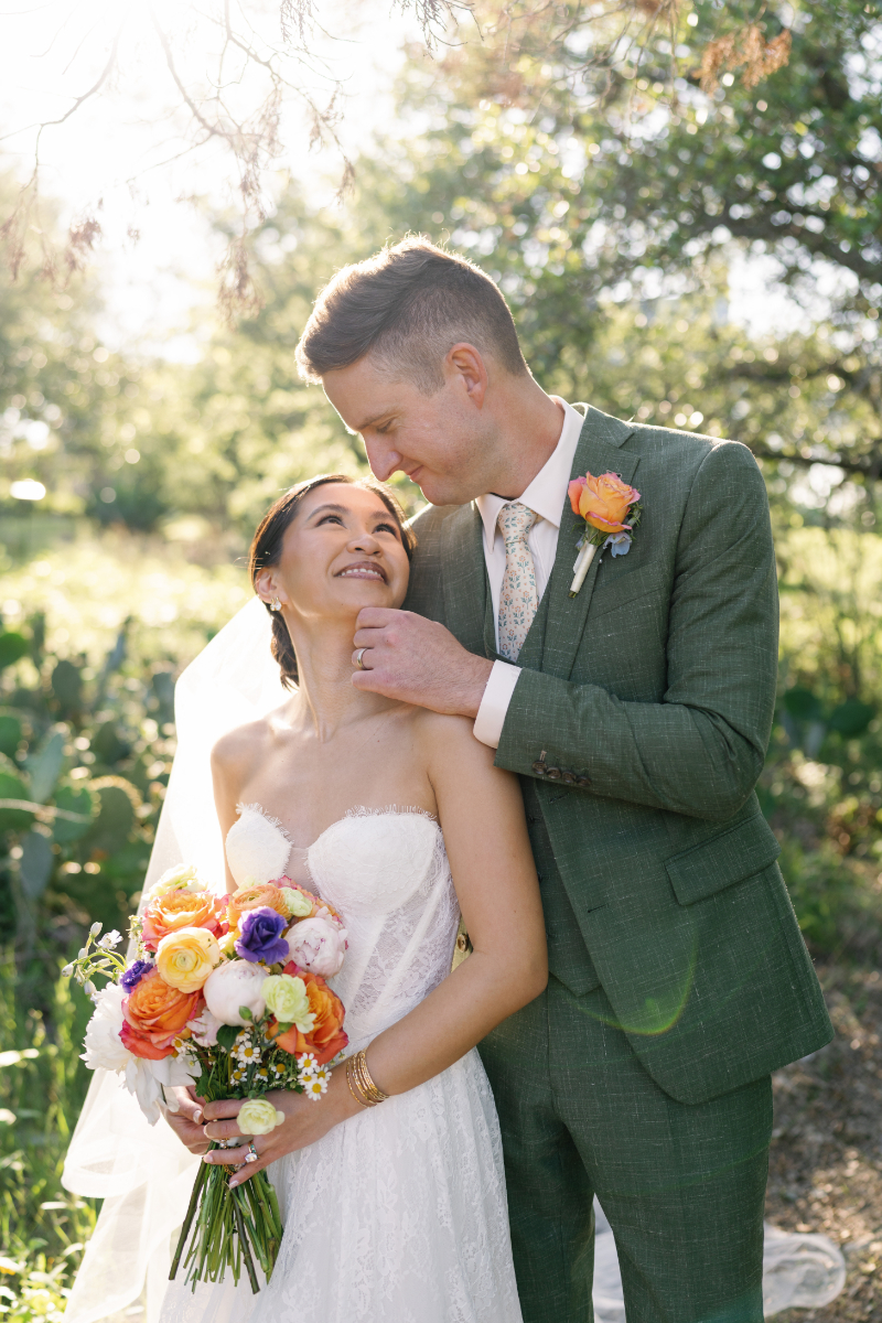A bride in a white dress holds a bouquet of colorful flowers while the groom in a green suit gently touches her chin outdoors in sunlight.