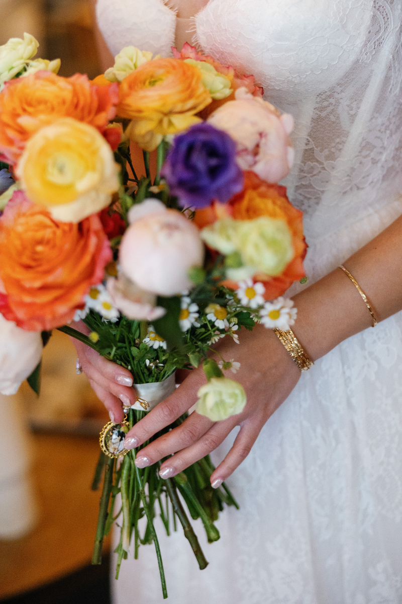 Person in a white lace dress holds a bouquet of colorful flowers, including orange, yellow, pink, and purple blooms. Gold bracelet and ring are visible on their left hand.