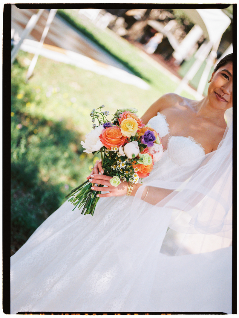 A bride in a white strapless dress holds a colorful bouquet of flowers outdoors, with part of her face and veil visible.