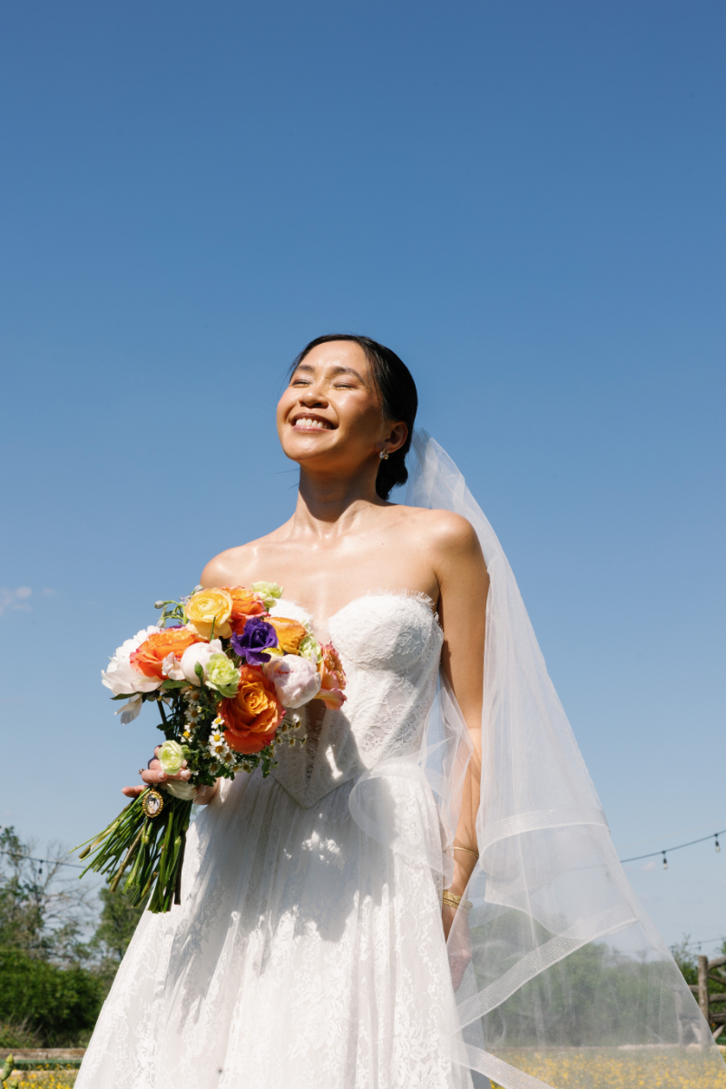 A bride in a white dress and veil holds a colorful bouquet and smiles outdoors under a clear blue sky.
