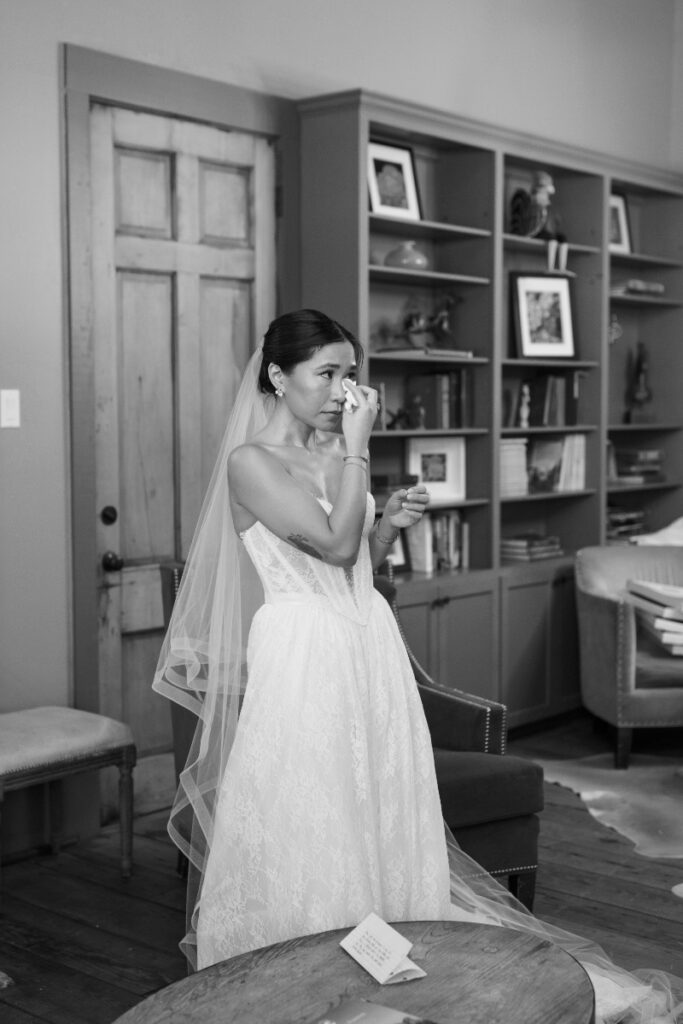 A bride in a white dress and veil wipes tears from her face while standing in a room with bookshelves and furniture.