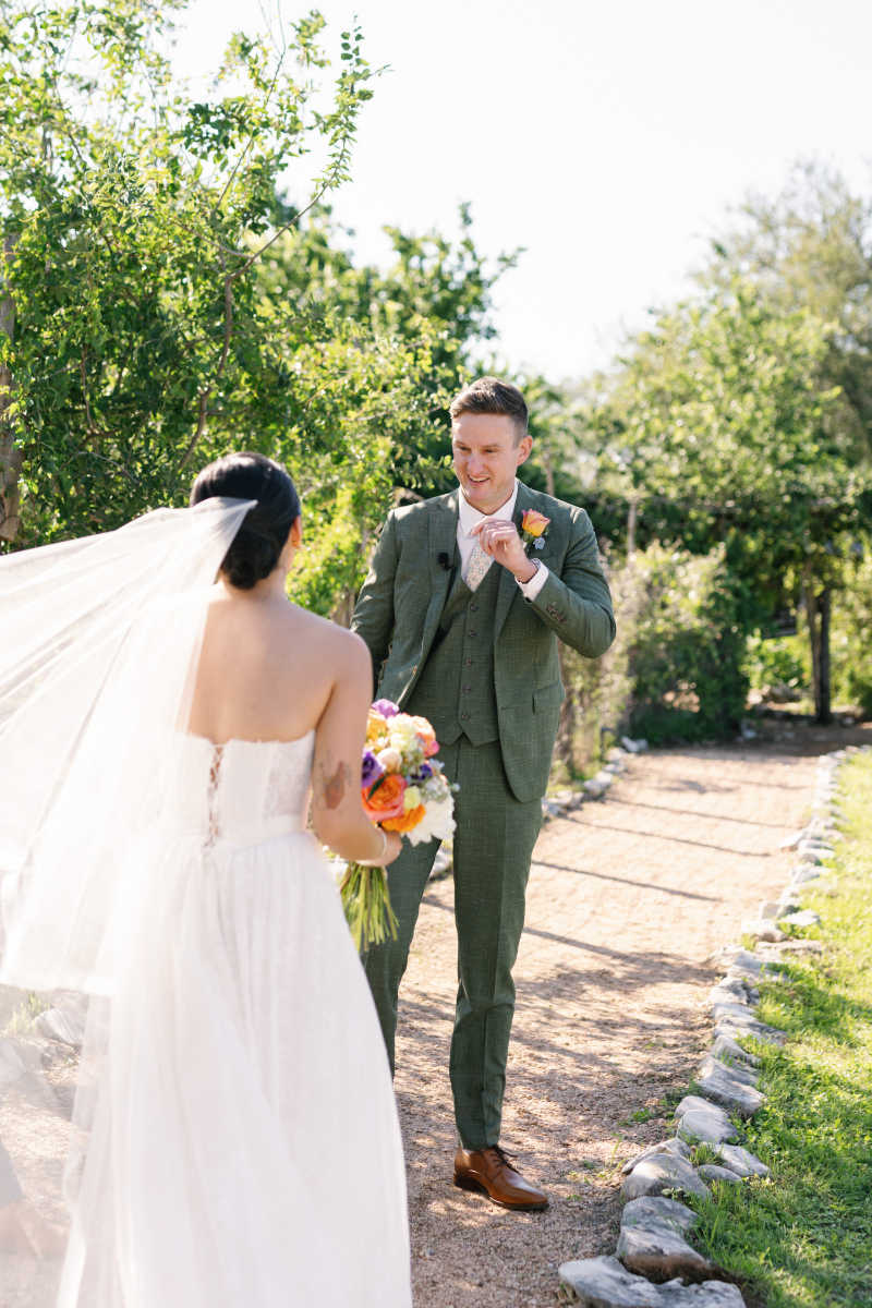A bride in a white dress and veil holding a bouquet faces a groom in a green suit on a sunlit garden path.