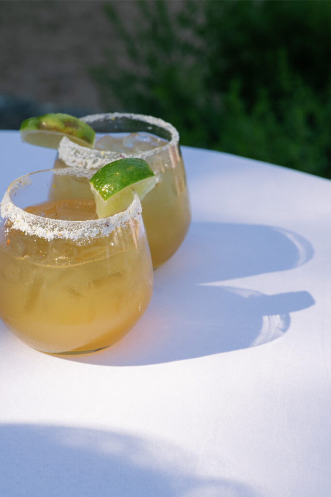 Two round glasses of iced cocktails with salted rims and lime wedges sit on a white table in sunlight, casting clear shadows.