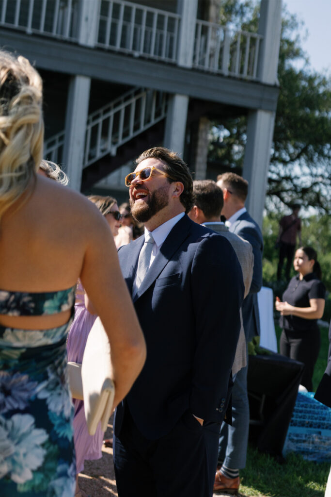 A man in a navy suit and sunglasses smiles while talking to others at an outdoor event near a building with white railings and stairs.