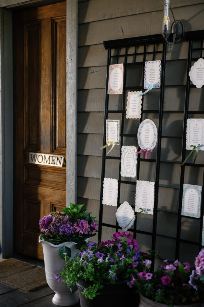 A wooden door labeled "WOMEN" is next to a trellis displaying cards with names, surrounded by potted purple and pink flowers.