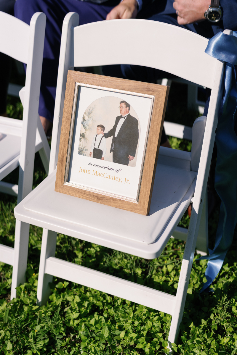 A framed memorial photo of an adult and child in formal wear sits on a white chair outdoors, with "In memoriam of John MacCauley, Jr." written below the image.