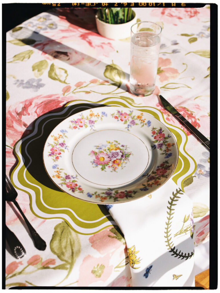 A floral-patterned plate sits on a green charger atop a flower-printed tablecloth, with cutlery, a napkin, and a glass of pink drink beside it.