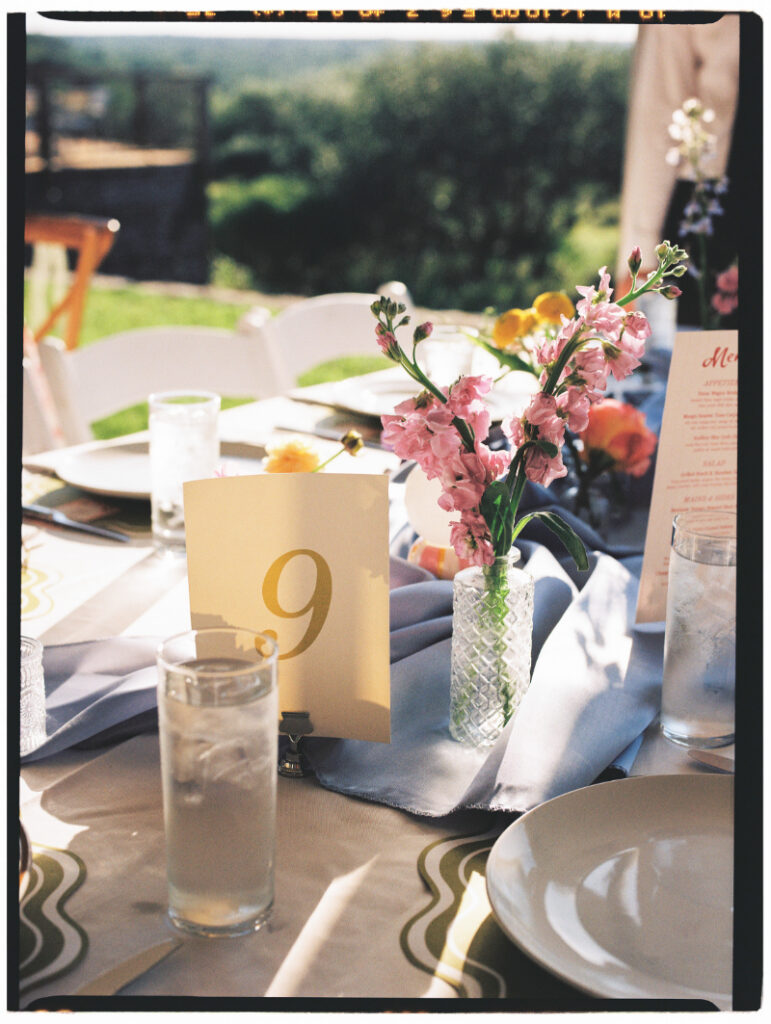 A table set for an event with a number 9 card, glassware, plates, menus, and a small vase of pink flowers on a patterned tablecloth.