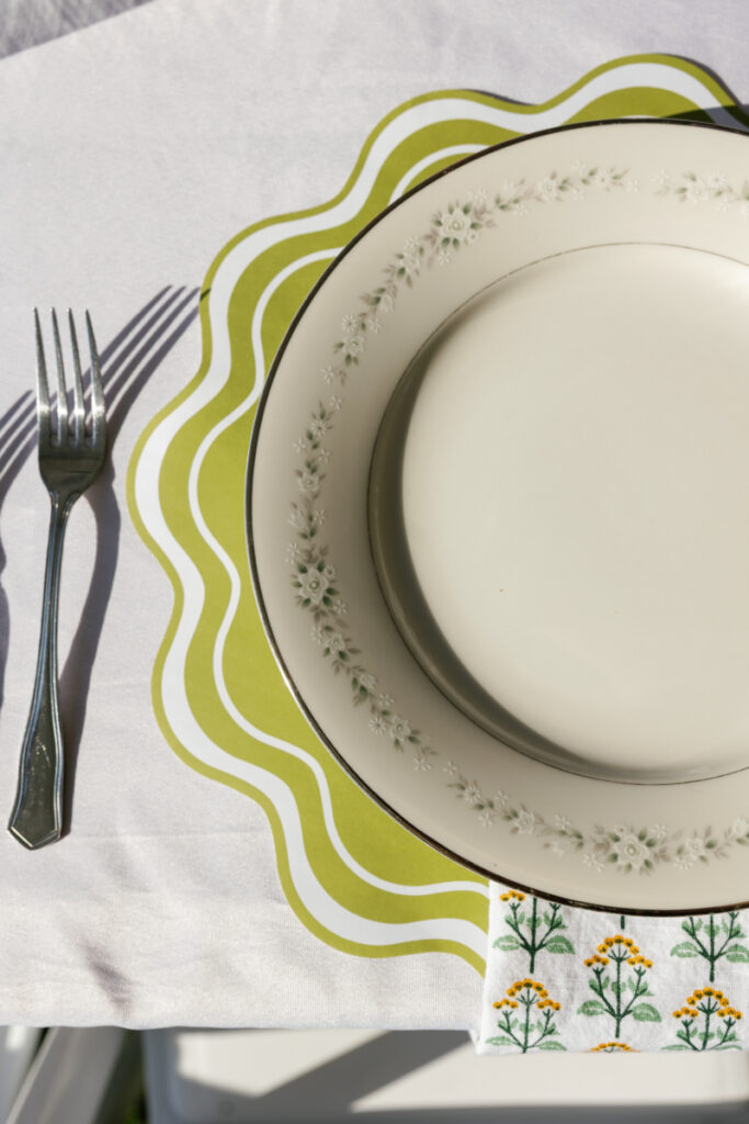 A white plate with a floral border sits on a green wavy placemat, next to a fork and a white napkin with yellow flower designs, all on a white tablecloth.